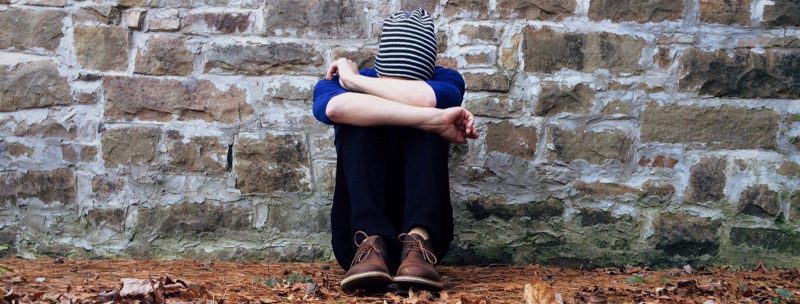 Depressed man with striped stocking cap on sitting on leaf covered ground with head on knees in front of a stone wall.