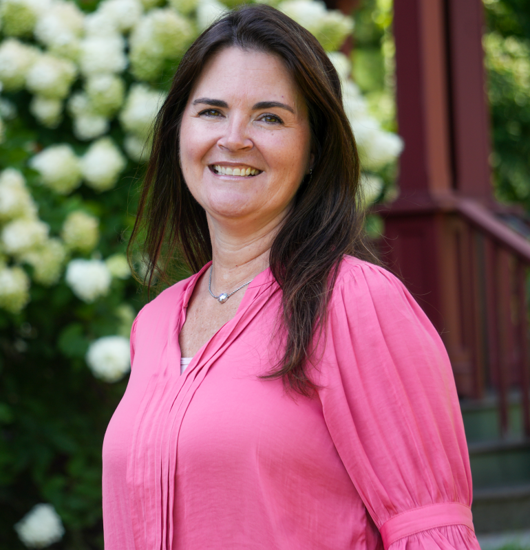 woman smiling while wearing a bright pink blouse, standing in front of a flowering bush. She has long dark hair.