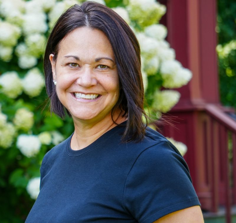 woman with dark hair smiling and standing in front of a flowering shrub.