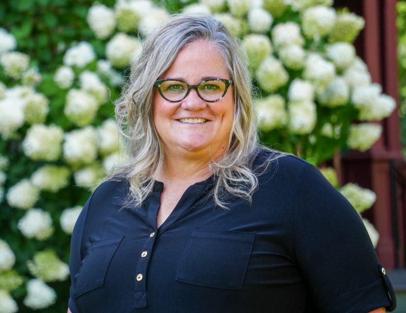 woman with long blonde hair and glasses smiling in front of flowering bush.