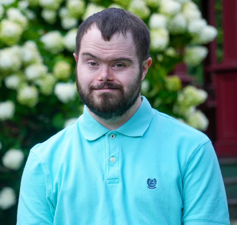 man with beard in a blue shirt, smiling in front of flowering bush.