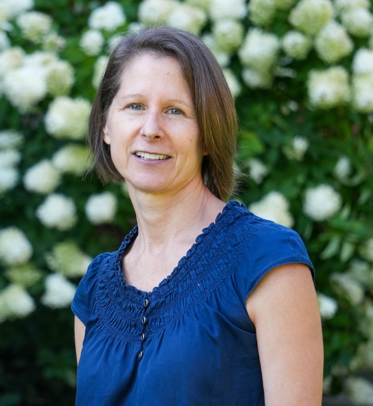 woman with short hair and blue dress smiling in front of flowering bush.
