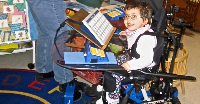 Image of a young girl with glasses in a wheelchair with a lap tray.  On the lap tray are a variety of alternative communication devices.